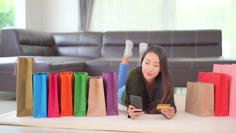 A-pretty-young-woman-lying-on-the-floor-surrounded-by-colorful-shopping-bags-double-checks-her-credit-card-number-with-her-smartphone-screen