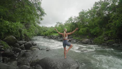 Woman-going-into-Tree-Pose-standing-on-rock-along-peaceful-stream-in-jungle