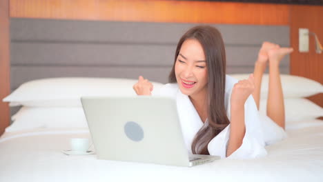 A-young-woman-in-a-terry-cloth-robe,-lying-on-her-stomach-on-a-comfortable-hotel-bed-works-on-her-laptop