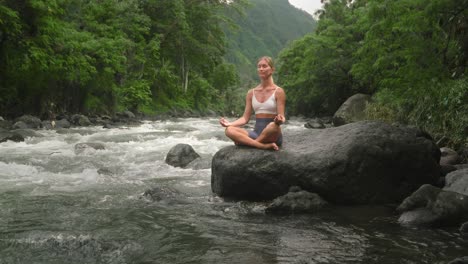 Young-female-traveler-outdoors-in-nature-relaxing-sounds,-easy-yoga-pose-on-rock