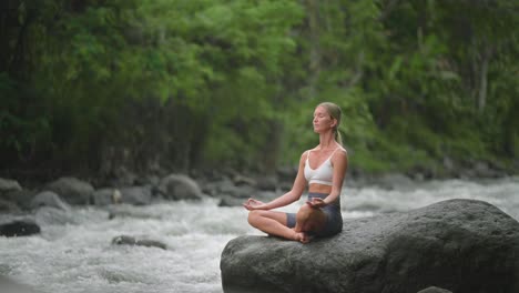 Pretty-caucasian-woman-practicing-Sukhasana-pose-on-rock-with-flowing-river