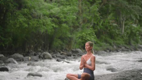 Female-meditating-alone-beside-tropical-river-in-Bali,-easy-pose,-Namaste