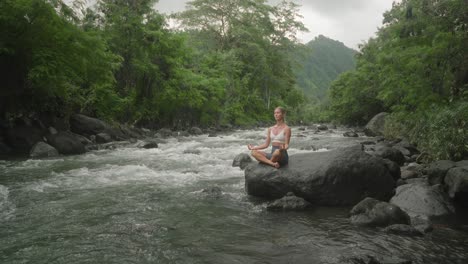 Soul-searching-attractive-female-in-lotus-pose-on-rock-along-calming-river