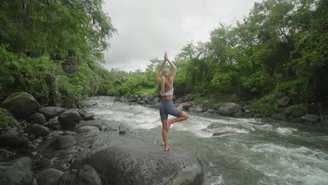 Slender-female-practicing-Tree-Pose-outdoor-next-to-jungle-river,-Vrksasana