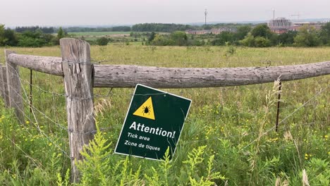 Tick-area-warning-sign-on-rural-park-trail-fence-with-tall-grass-in-farmland-field
