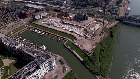 Backwards-aerial-movement-showing-construction-site-of-luxury-apartment-building-at-river-IJssel-riverbed-revealing-the-wider-urban-cityscape-with-skyline-of-tower-town-Zutphen-in-the-background