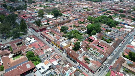 Drone-aerial-footage-of-car-and-motorbike-street-traffic-in-Antigua,-Guatemala-colonial-town-showing-bright-colorful-red-rooftops,-and-green-tree-tops