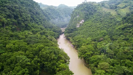 Drone-aerial-panorama-of-flowing-rainforest-river-valley-lined-with-lush-green-trees-surrounded-by-dense-jungle-hillsides-and-mountains