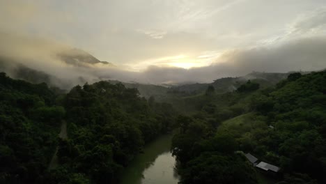 Drone-aerial-footage-of-dusk-sunset-over-misty-and-cloudy-rainforest-valley-and-river-lined-with-lush-dark-green-trees-near-Semuc-Champey-National-Park,-Guatemala