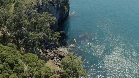 Kayakers-floating-along-steep-cliff-shore-of-island,-adventure-activity-in-New-Zealand,-bright-sunny-day