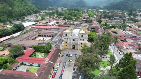 Drone-footage-of-Iglesia-de-la-Merced-Church-in-Antigua,-Guatemala-on-a-bright-cloudy-day-with-colorful-red-town-rooftops-and-green-forest-surrounding-the-city