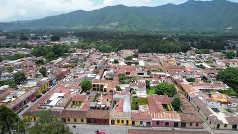 Drone-aerial-footage-of-city-block-in-Antigua,-Guatemala-colonial-town-showing-bright-and-colorful-red-rooftops-and-green-tree-tops-and-forest-mountain-in-the-background