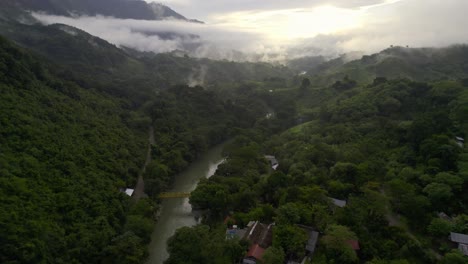 Drone-aerial-footage-of-sunset-over-misty-and-cloudy-rainforest-valley-and-river-lined-with-lush-dark-green-trees-near-Semuc-Champey-National-Park,-Guatemala-surrounded-by-hillsides-and-mountains