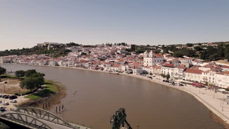 Alcacer-Do-Sal-Brücke-Gegen-Stadtbild-Am-Flussufer,-Portugal,-Fliegen-über-Den-Fluss-Sado