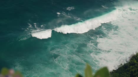 Large-wave-breaks-at-tropical-shore-of-Bali,-seen-from-top-of-cliff,-blue-water