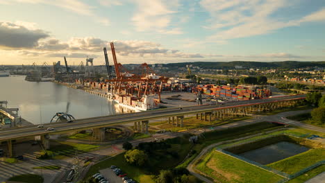 Cars-driving-on-highway-beside-Shipyard-with-cranes-and-container-in-Gdynia,Poland---Beautiful-golden-sunrise-in-the-morning---Aerial-backwards-flight