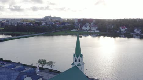Flying-towards-church-bell-tower-in-Reykjavik-with-view-of-lake,-bright-sunlight