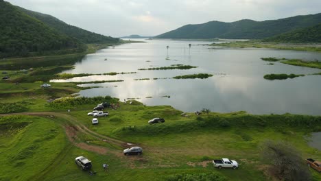 Aerial-footage-revealing-an-outdoor-adventure-scene-in-which-trucks-and-cars-parked-at-the-lake,-people-fishing,-picnic,-fantastic-landscape-and-lake-reflecting-clouds-and-sunset-in-Thailand