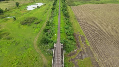 Aerial-footage-over-an-elevated-railway-revealing-a-farm-on-the-right,-grassland-on-the-left-and-a-ponds,-in-Saraburi,-Thailand