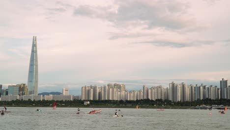 Lotte-Tower-In-South-Korea---Han-River-With-People-Enjoying-Windsurfing,-Paddleboarding,-Boating,-And-Kayaking-With-Seoul-Skyline-In-The-Background