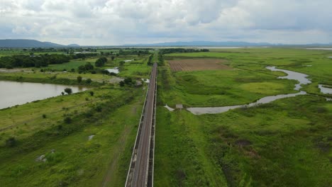 Aerial-footage-towards-a-scenic-horizon-revealing-a-lake-on-the-left,-wetlands,-farmlands,-elevated-railway-with-a-gorgeous-landscape-and-lake,-in-Saraburi,-Thailand
