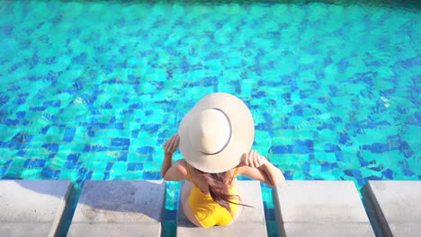 Top-View-of-Petite-Female-in-Yellow-Swimsuit-and-Floppy-Hat-Sitting-on-Poolside-at-Tropical-Destination-on-Sunny-Day,-Full-Frame