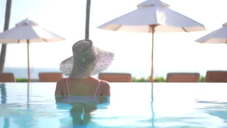 Dreamy-Scene,-Back-View-of-Female-With-Summer-Hat-in-Swimming-Pool-on-Sunny-Day-Looking-at-Poolside-and-Skyline,-Full-Frame-Slow-Motion