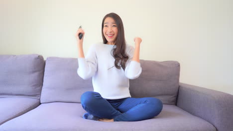Happy-Asian-Woman-Watching-News-on-TV-Holding-Remote-and-Celebrating-With-Hands-Gesture,-Lottery-or-Sport-Winning,-Full-Frame-Slow-Motion