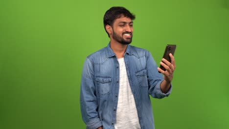 Attractive-Asian-man-talking-on-a-mobile-phone-and-smiling-in-front-of-green-screen:-hand-waving-hand-to-a-known-person-on-phone