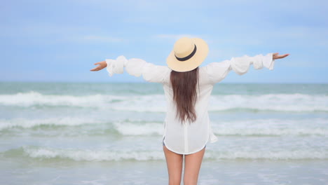 Joyful-woman-with-long-hair-and-floppy-hat-raising-and-spreading-hands-in-front-of-waves-and-horizon-of-tropical-sea,-back-view,-full-frame,-slow-motion