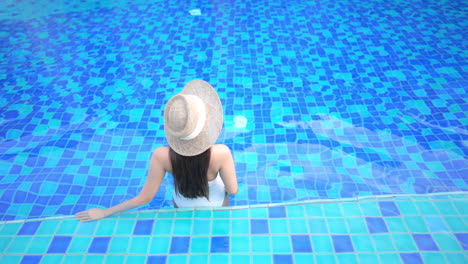 Back-View-of-Woman-in-Swimsuit-and-Floppy-Hat-Standing-Alone-in-Blue-Swimming-Pool,-Full-Frame
