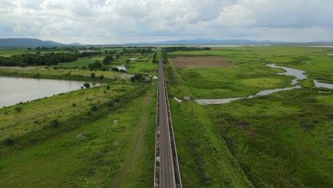 Aerial-reverse-footage-revealing-an-elevated-railway-through-wetlands-and-farmlands,-gorgeous-landscape-and-lake,-in-Saraburi,-Thailand