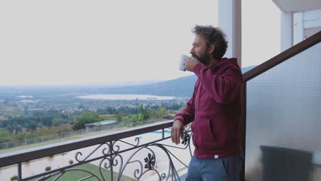 Mann,-Der-Vom-Stuhl-Aufsteht-Und-Auf-Einem-Balkon-Mit-Atemberaubender-Aussicht-Auf-Den-Horizont-Blickt,-Während-Er-Eine-Tasse-Kaffee-Hält