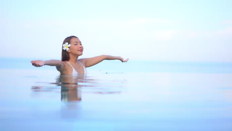 A-young-woman,-chest-high-in-the-water-of-an-infinity-edge-pool,-raises-her-arms-from-the-water-to-the-sky-and-smiles