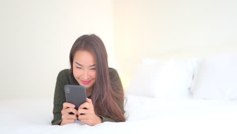 A-pretty-young-woman-lying-on-a-big-comfortable-bed-smiles-as-she-inputs-text-into-her-smartphone