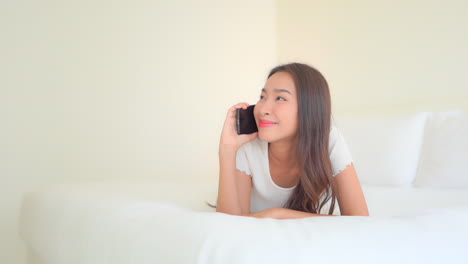 A-pretty-young-woman-lying-on-a-comfortable-hotel-suite-bed-talking-on-her-smartphone-and-reacting-to-what-she-hears