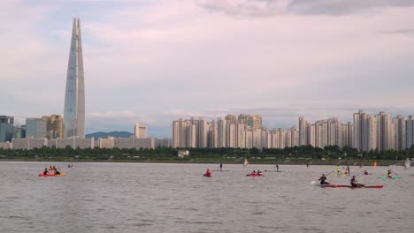 Gente-Disfrutando-Del-Río-Hangang-Recreativo---Windsurf,-Paddle-Surf-Y-Kayak-Con-El-Horizonte-De-La-Ciudad-De-Seúl-En-El-Fondo,-Lotter-World-Tower