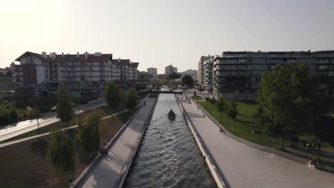 Water-canal-in-Aveiro-city-with-buildings-in-horizon-and-tourist-canoe,-aerial-view
