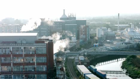 Industrial-river-chemical-warehouse-building-with-factory-steam-emissions-in-early-morning-sunlight-aerial-descending