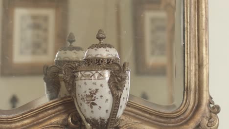Close-up-of-vase-with-delicate-ornaments-in-front-of-ancient-mirror-with-18th-century-frame