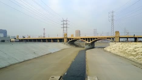 Downtown-LA-river-historic-urban-flood-control-concrete-water-channel-towards-urban-viaduct-bridge,-aerial-view