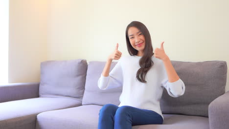 An-Asian-Thai-Woman-Showing-Big-Thumbs-up-Gesture-while-Sitting-on-the-Couch-in-Casual-Clothes-at-Home-Interior