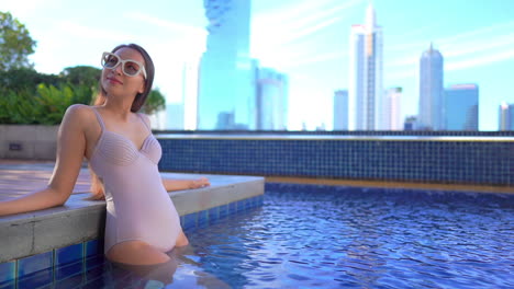An-attractive-young-woman-in-a-one-piece-swimsuit-leans-along-the-side-of-a-pool-with-a-modern-city-skyline-behind-her