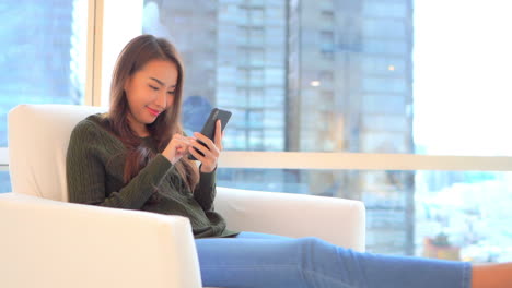 A-young-woman-sits-comfortably-in-an-easy-chair-backed-by-modern-skyscrapers-in-the-window-as-she-taps,-scrolls,-and-interacts-with-her-smartphone