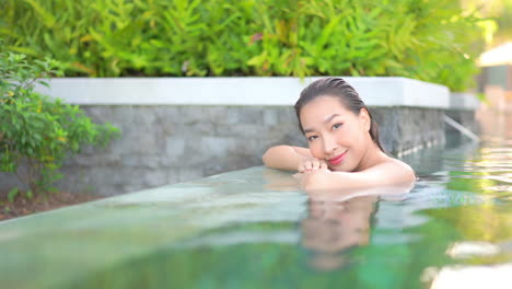 Close-up-of-a-pretty-young-woman-in-a-swimming-pool,-leaning-along-the-edge-surrounded-by-lush-tropical-plants