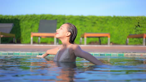 Asian-woman-in-swimwear-turns-around-with-spread-on-sides-hands-and-closed-eyes-inside-swimming-pool-at-a-private-resort-in-Spain,-slow-motion-daytime-sunny