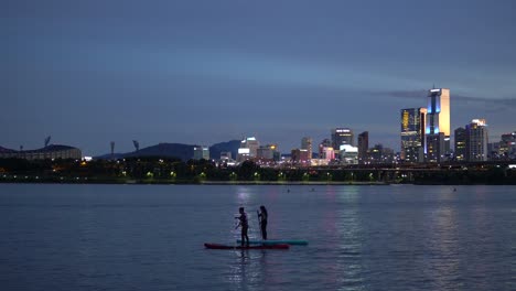 Silhouette-of-Two-People-Stand-Up-Paddleboarding-on-Han-River-at-Twilight,-Cheongdam-Bridge-with-Night-Cars-Traffic-and-Jamshil-District-of-Seoul-on-Colorful-Purple-Sky-Background,-Static-4k