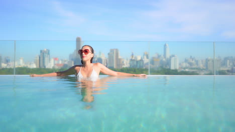 Sexy-lady-leaning-on-the-edge-of-a-rooftop-infinity-pool,-a-beautiful-young-woman-enjoys-the-sun-and-water-with-Bangkok-city-skyline-behind-her,-Thailand-urban-vacation