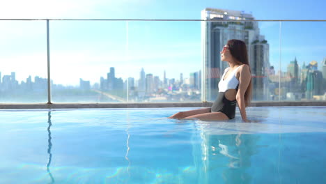 Wunderschöne-Asiatische-Frau,-Die-Im-Schwimmbad-Auf-Dem-Dach-Mit-Atemberaubender-Skyline-Von-Bangkok-Sitzt