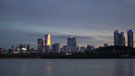 Nacht-Seoul-Vom-Flussufer-Des-Han-flusses-Bei-Farbenprächtigem-Sonnenuntergang,-Landschaft-Mit-Wolkenkratzern-Wie-Glasturm,-Handelsturm-Und-Asem-turm-Unter-Sonnenlicht,-Yacht-Vorbeifahren,-Wenn-Menschen-Auf-Dem-Fluss-Paddeln